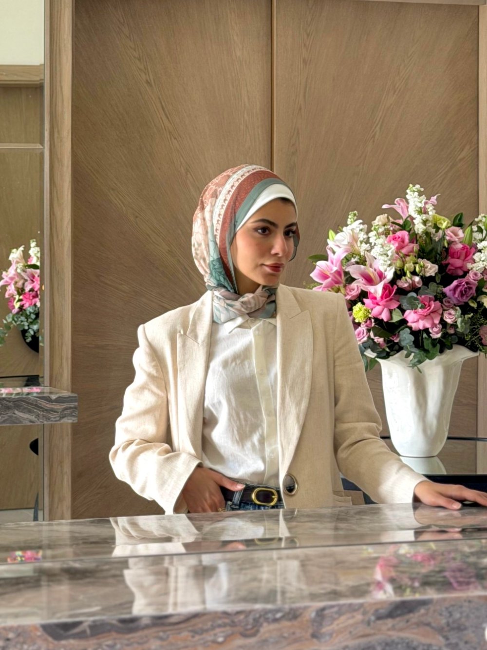 Farah Sherzman in a cream linen blazer, standing at a marble counter beside a bouquet of pink lilies and roses